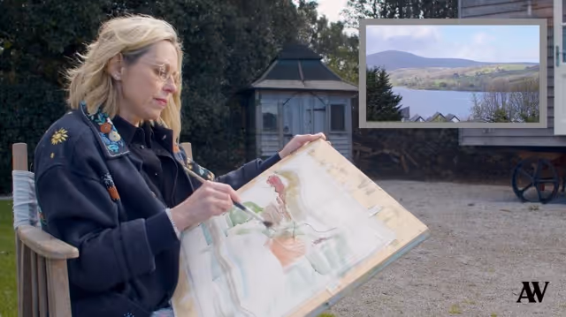 Woman outdoors painting on a board with a scenic view of hills and a lake in an inset frame.