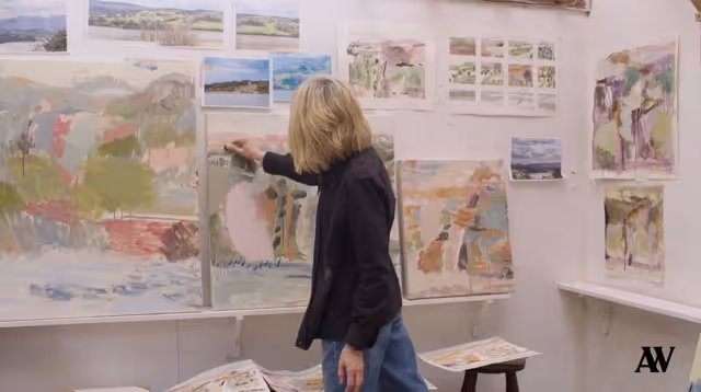 Artist in a studio painting on a canvas surrounded by landscape artworks on walls.