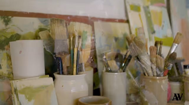 Various paintbrushes held in jars with paintings blurred in the background.