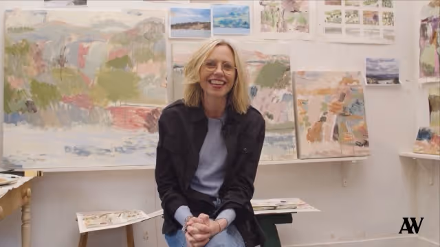 Smiling woman with glasses sitting in front of abstract paintings in an art studio.