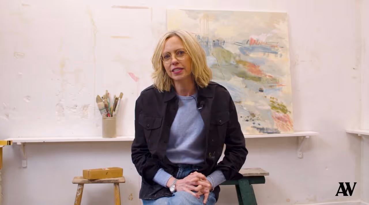 Blonde woman with glasses sitting in art studio with paintbrushes and abstract painting behind her.