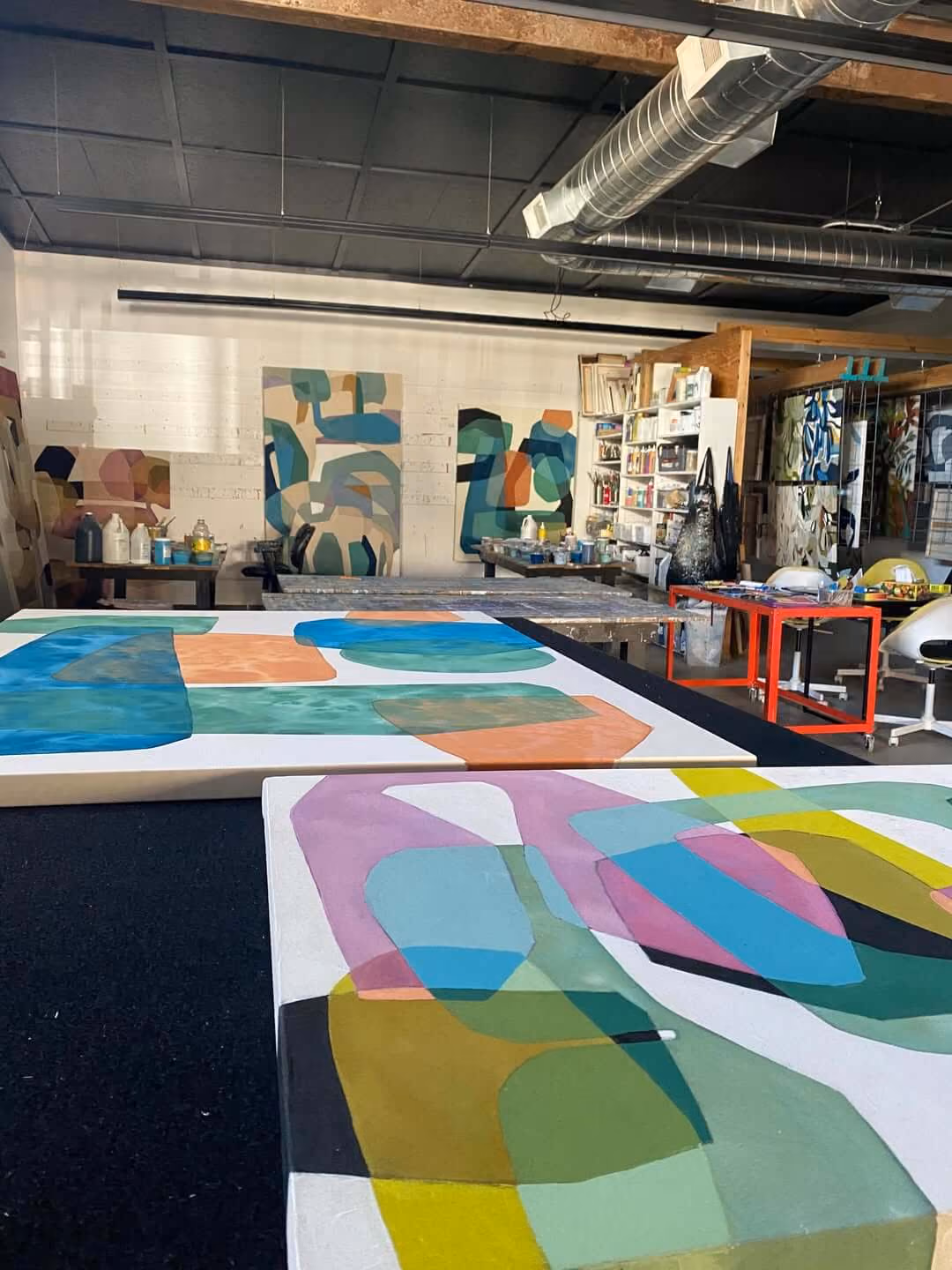 Art studio with colorful abstract paintings on tables and walls amid shelves and art supplies.