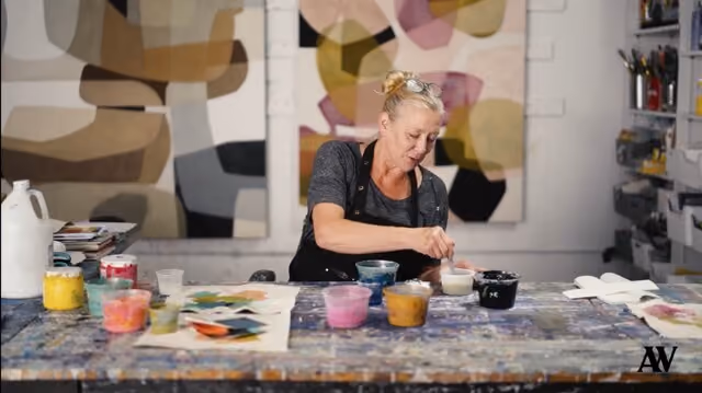 Woman artist mixing paint cups on a messy table with abstract paintings in the background.