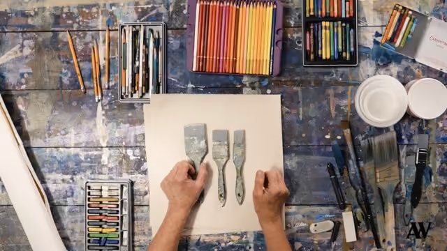 Hands holding three metal palette knives on a paint-splattered wooden table with pencils and brushes.
