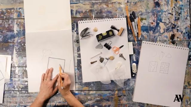 Hands drawing on sketchbooks amid art supplies scattered on a colorful, paint-splattered table.