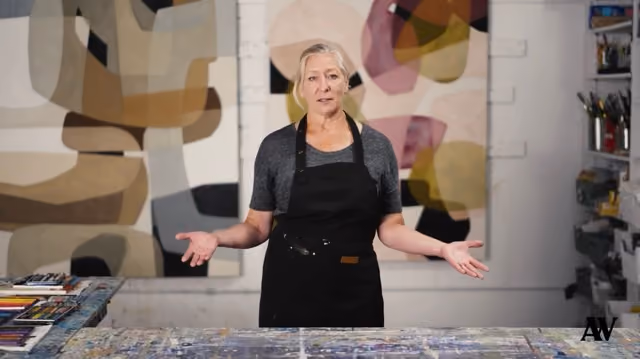 Woman artist in black apron standing behind a paint-covered table in a studio with abstract art on walls.