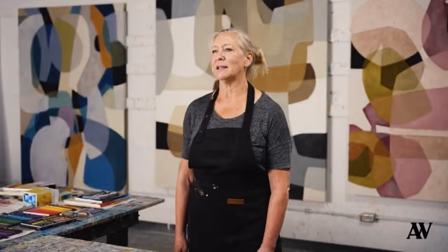 Woman artist in black apron standing in studio with abstract paintings on the walls behind her.