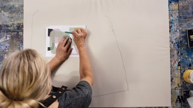 Person sketching a large outline on tan paper using a small reference image on a paint-splattered surface.