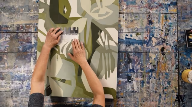 Person places a label on a green and beige abstract painting on a paint-splattered surface.