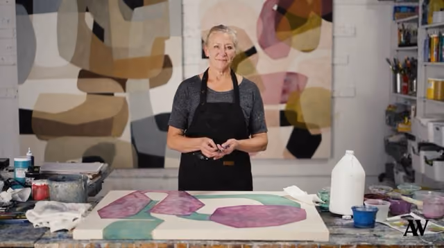 Woman artist in apron working on abstract painting with purple and green shapes in studio.