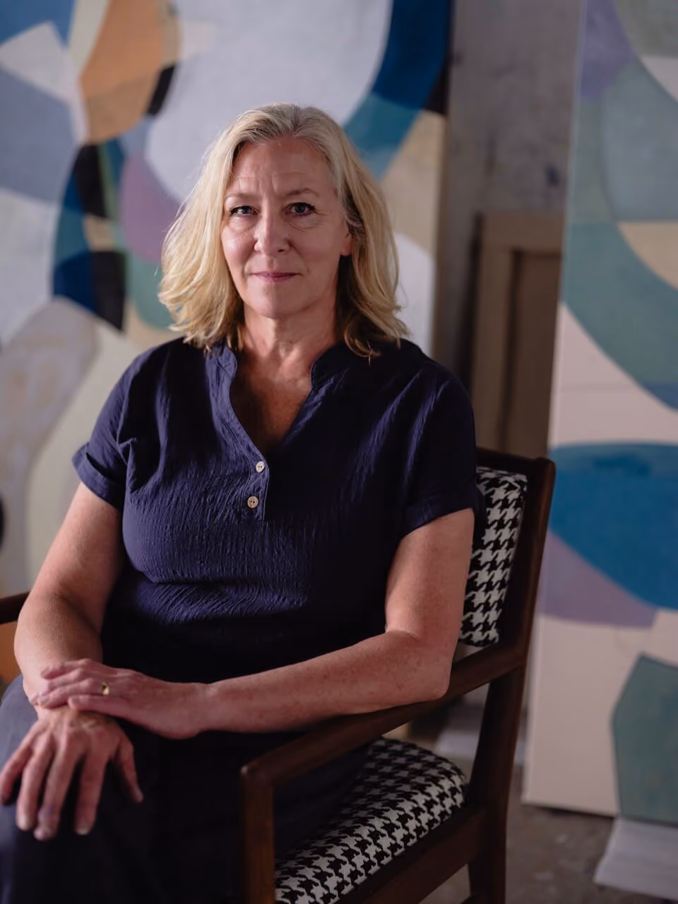 Middle-aged woman with blonde hair sitting on patterned chair in front of abstract colorful artwork.