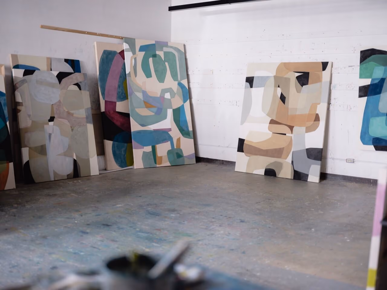 Abstract paintings with geometric shapes and muted colors leaning against a white wall in a studio.