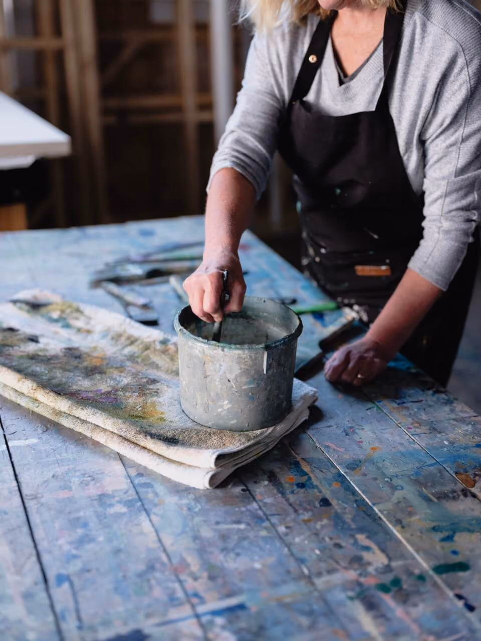 Person stirring paint in a bucket on a paint-splattered table with cloth underneath.