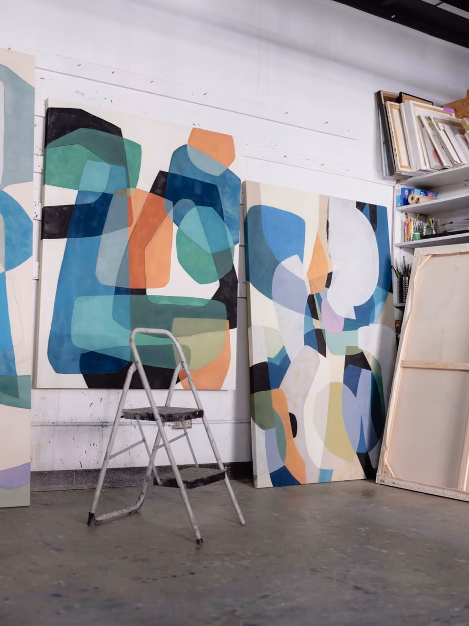 Art studio with abstract paintings in blue, green, orange, and white leaning against wall and a metal step ladder.
