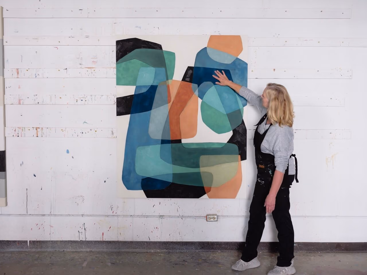 Artist in black apron touching a large abstract painting with layered blue, green, orange, and black shapes.