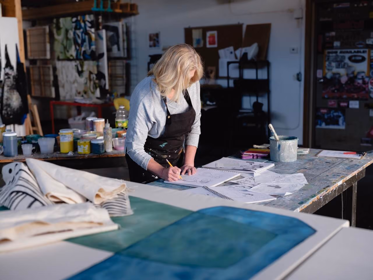 Woman with blonde hair drawing sketches at a cluttered art studio table with paints and papers.