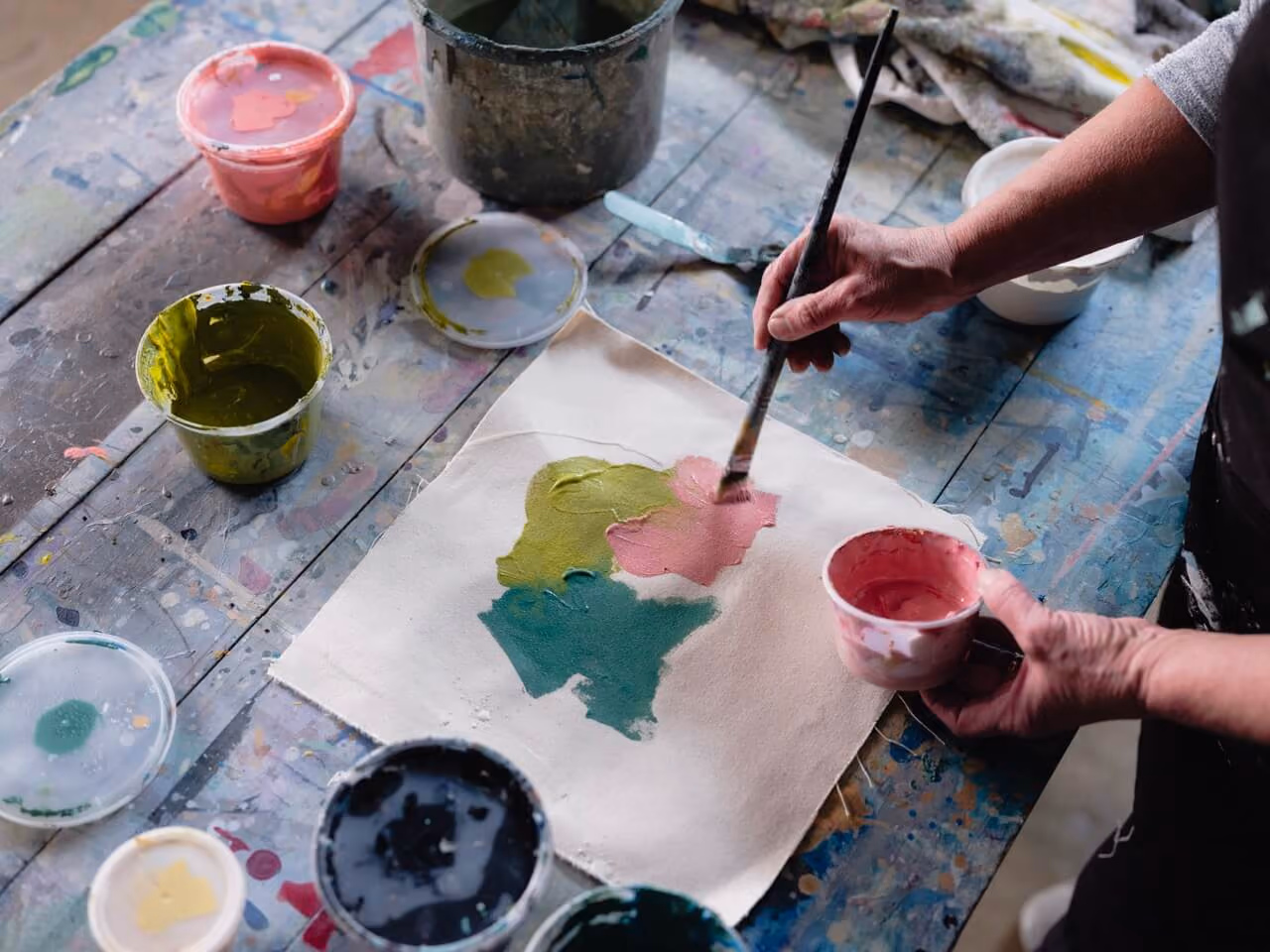 Person painting pink, green, and teal blobs on fabric with a brush over a paint-splattered table.
