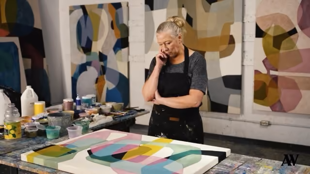 Woman artist in apron thoughtfully observing her colorful abstract painting in a studio workspace.