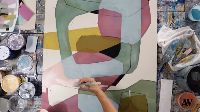 Artist painting abstract overlapping shapes in muted colors on paper with brushes and paint nearby.