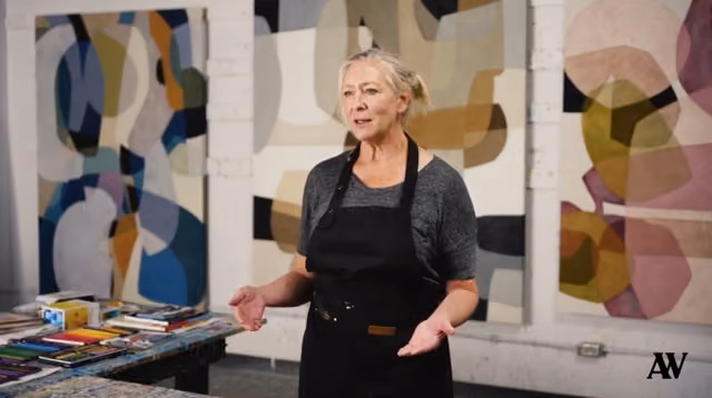 Woman wearing black apron stands in front of abstract paintings in an art studio.