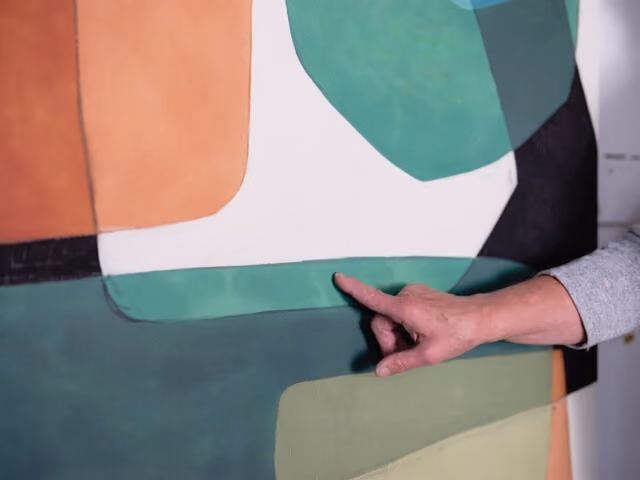 Hand pointing to an abstract painting with overlapping geometric shapes in green, black, white, orange, and beige.