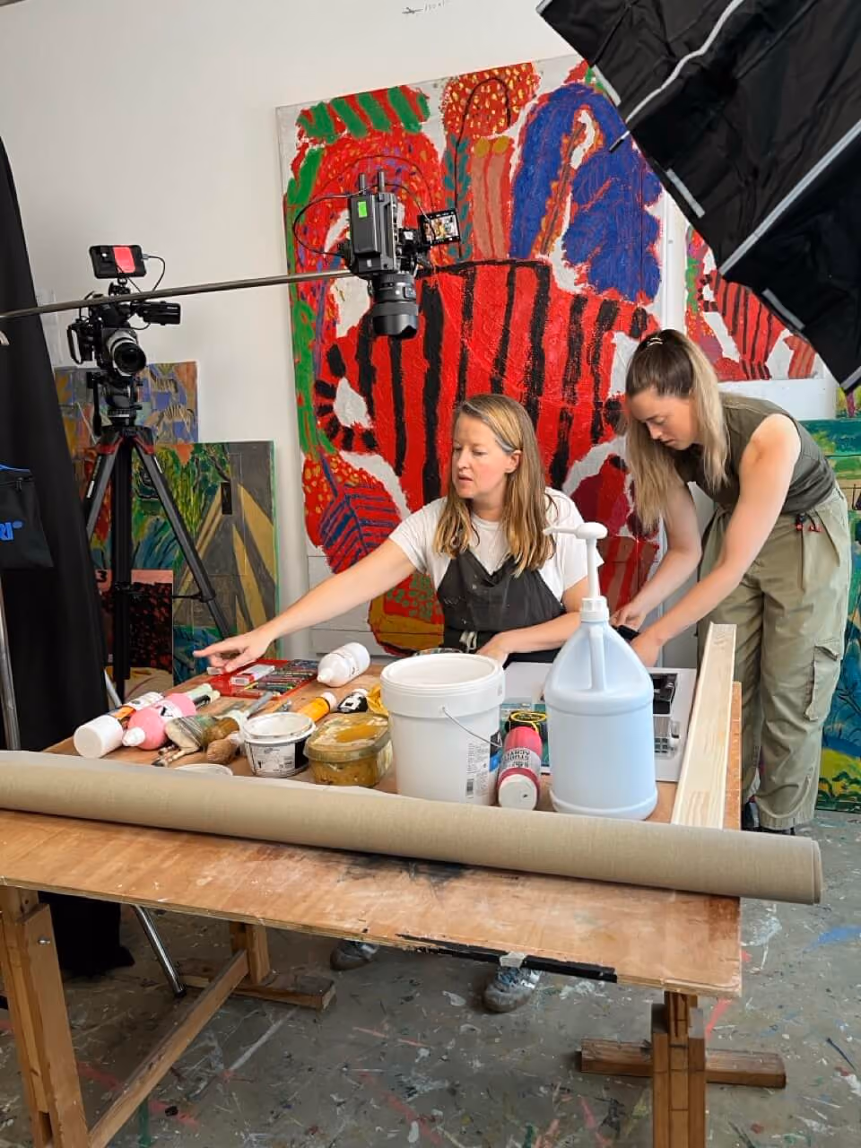Two women working with art supplies on a table in a studio with colorful paintings and cameras around.