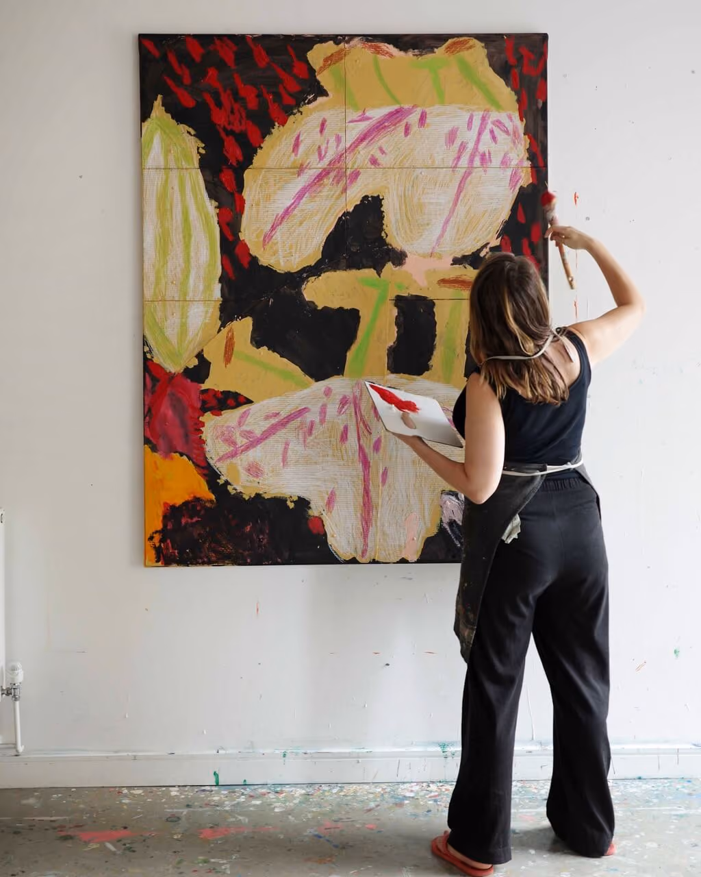 Woman in black apron painting a large abstract colorful artwork on a white wall in a studio.