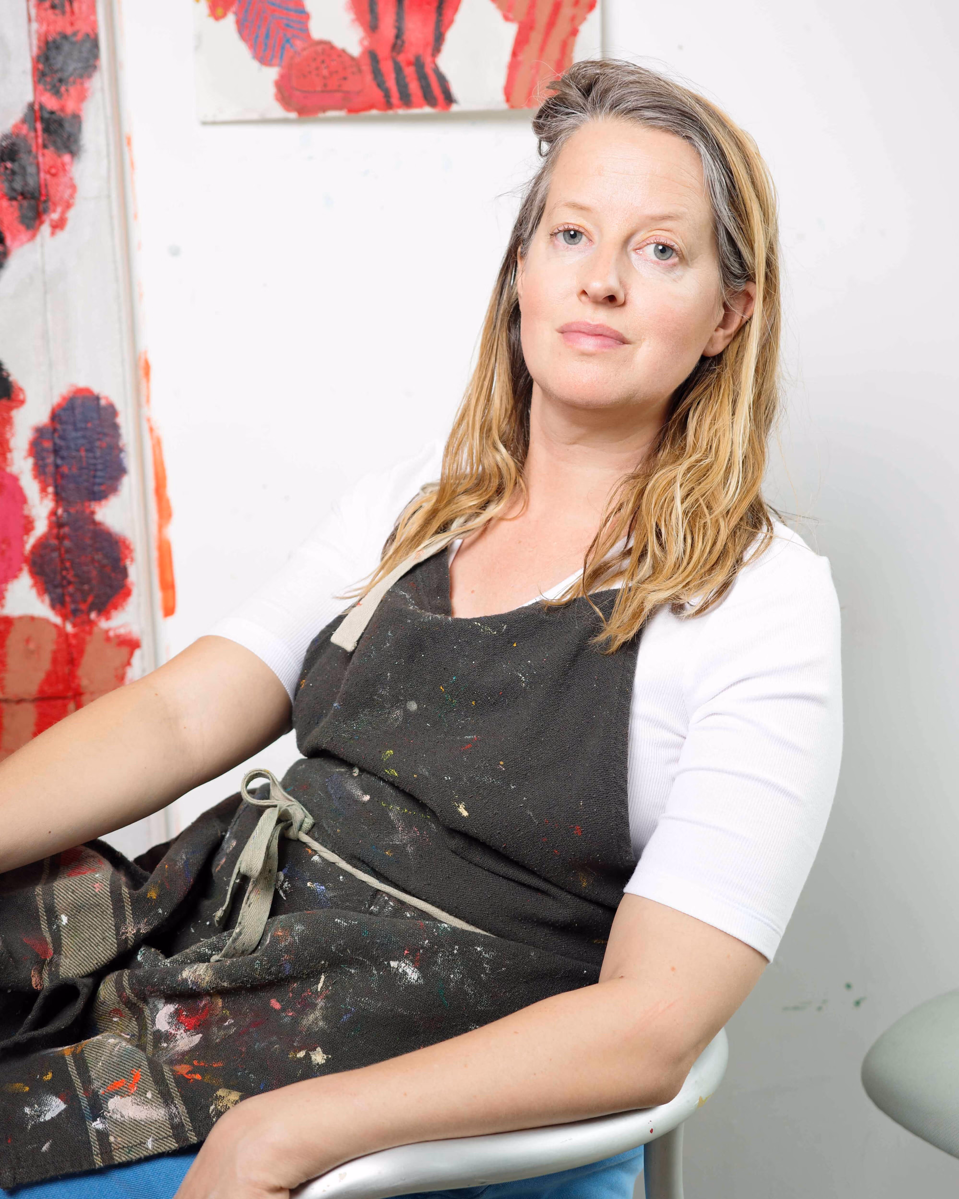 Woman in paint-stained apron sitting on chair against white wall with red artwork.