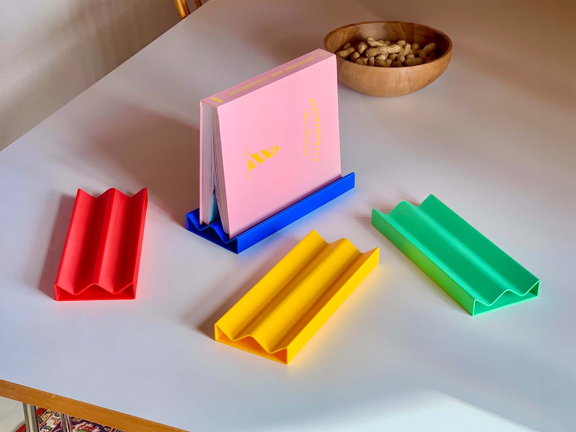 Four colorful plastic book or tablet holders in red, blue, yellow, and green on a white table, with a pink book placed in the blue holder and a wooden bowl of peanuts in the background.