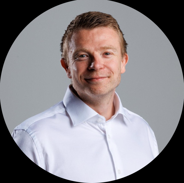 Smiling man with short brown hair wearing a white button-up shirt against a gray background.