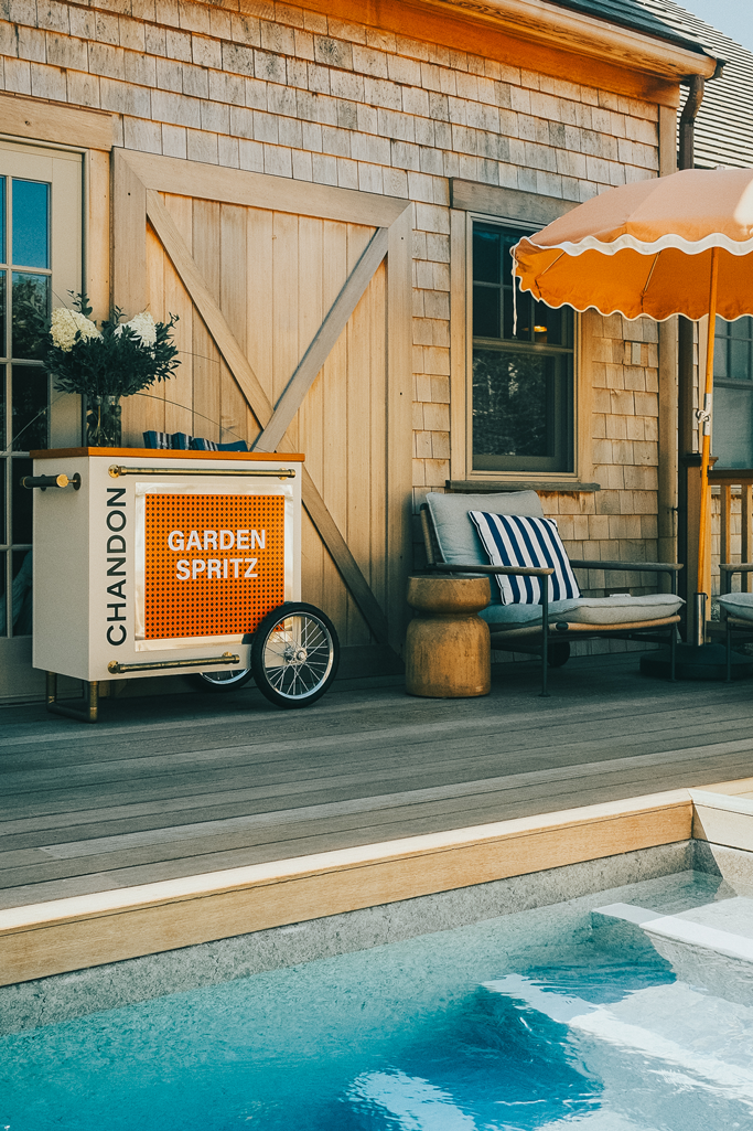 Outdoor wooden deck with a pool, a Chandon Garden Spritz cart, cushioned chairs with striped pillows, and an orange umbrella.