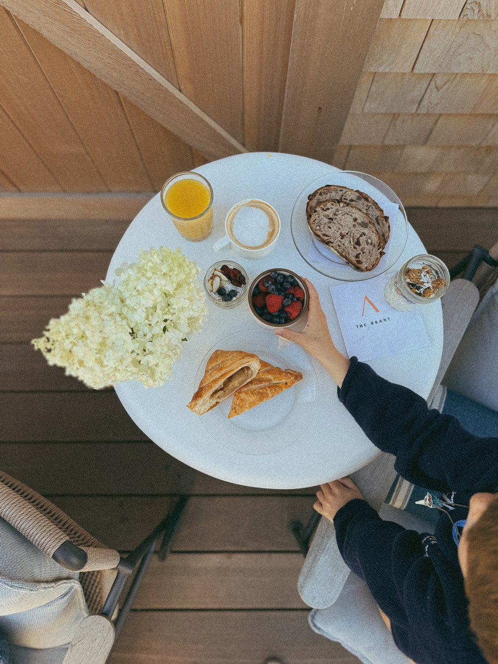 Top view of a small round white table with orange juice, latte, granola jar, two slices of nut bread, pastry triangles, a bowl of mixed berries, white flowers, and a person reaching for the bowl of berries.