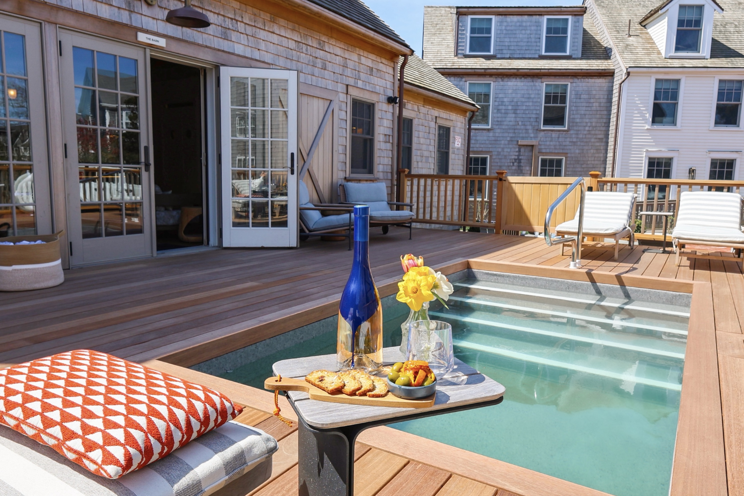 Poolside deck with wooden flooring, lounge chairs with cushions, a small table holding a blue bottle, glass, flowers, crackers, and olives under a blue sky.