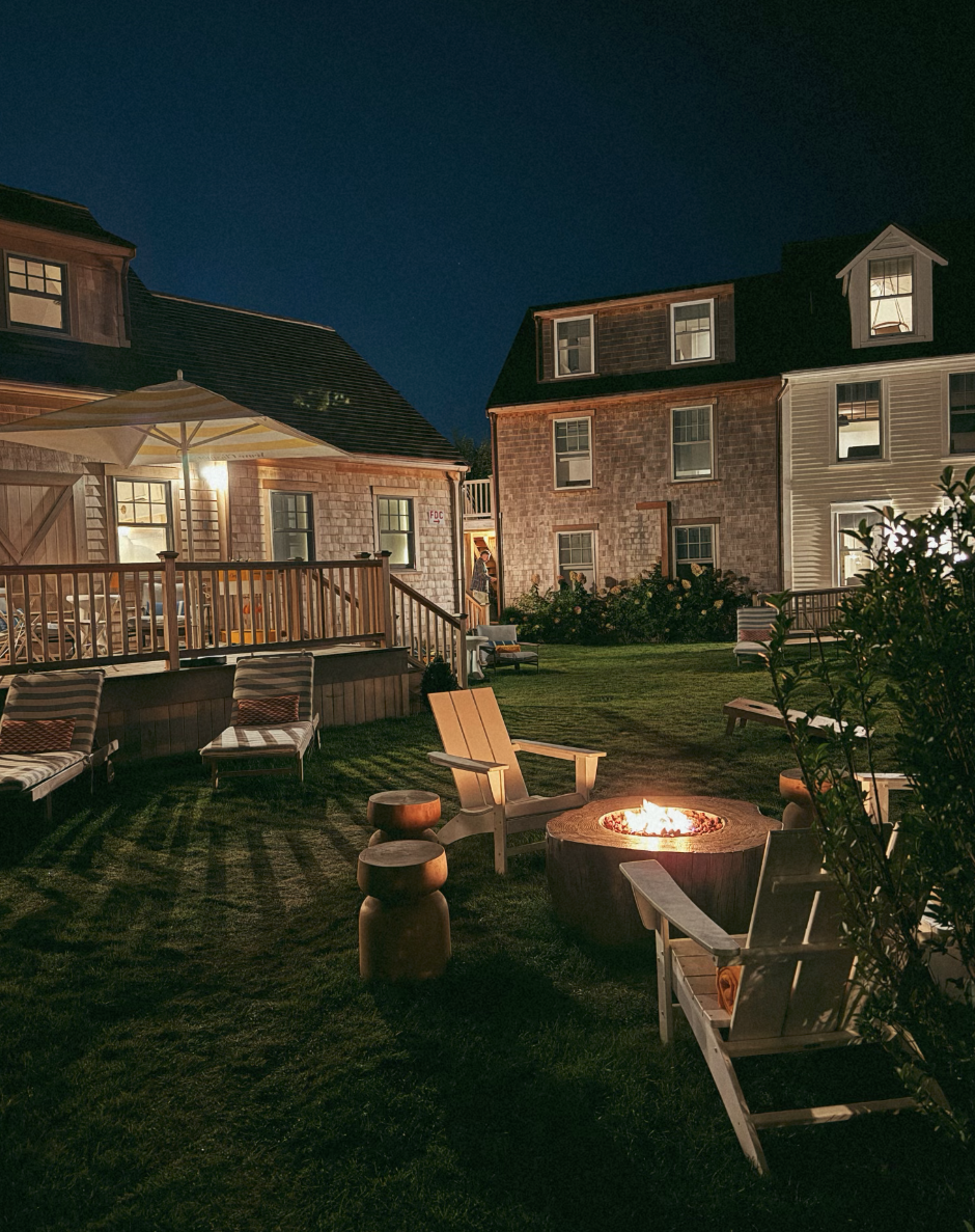Nighttime backyard scene with lit fire pit surrounded by wooden chairs and stools, adjacent to houses with illuminated windows and porch.