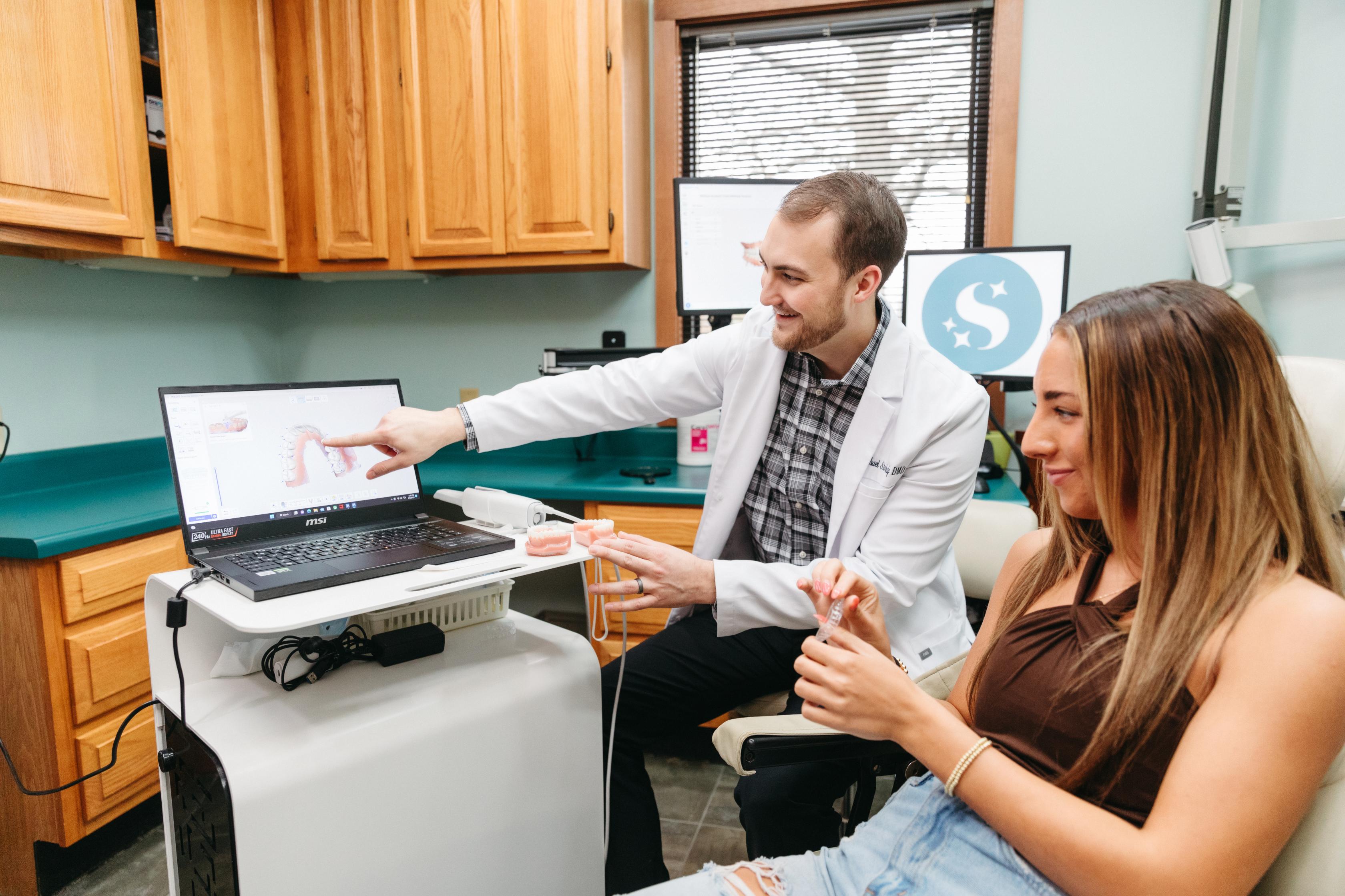 A smiling dentist in a white coat shows a 3D dental scan on a laptop to a female patient, who is holding a dental model in a modern exam room with wooden cabinets and green counters.