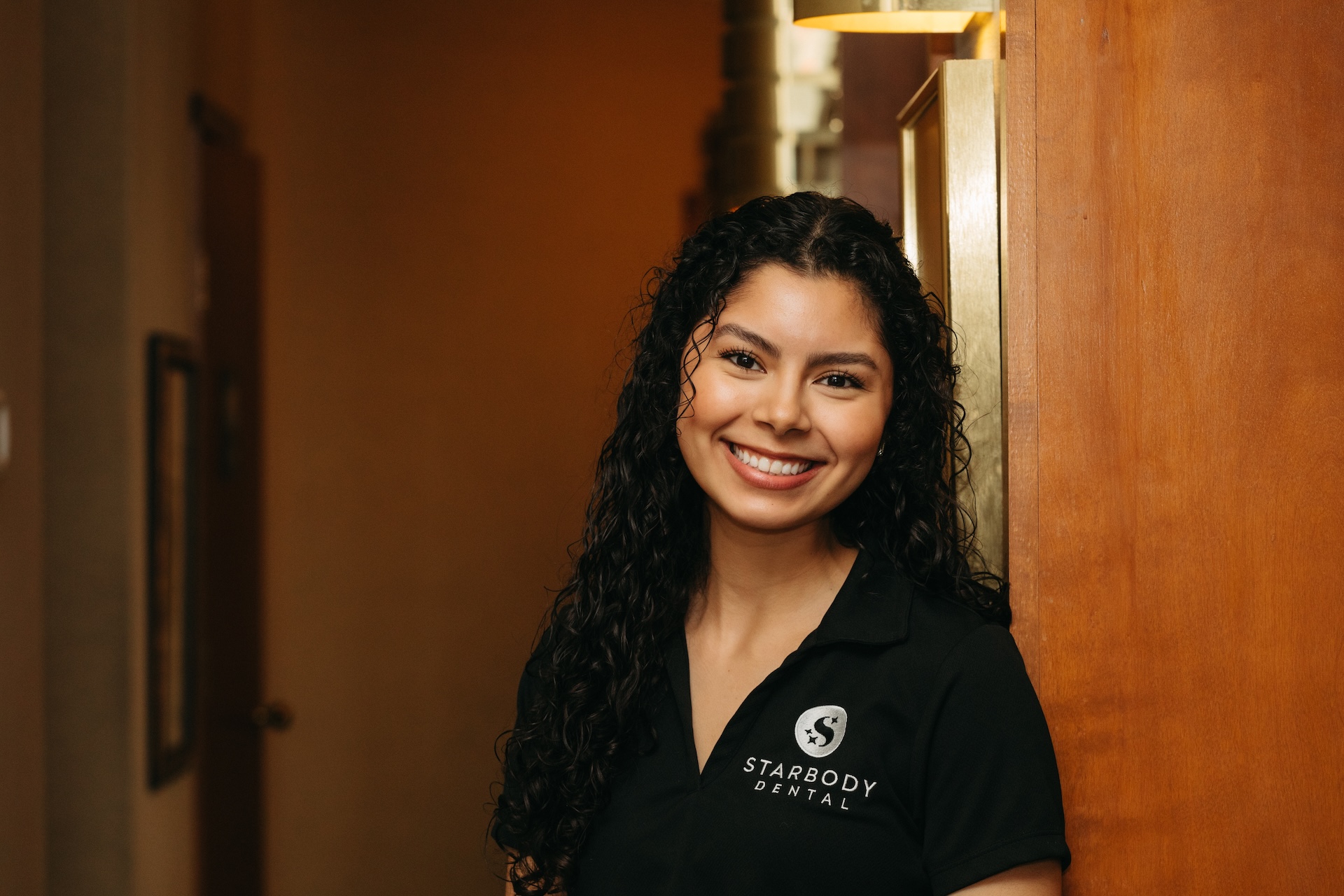 Smiling dental staff member in a black Starbody Dental uniform standing by a wooden wall, looking friendly and welcoming.