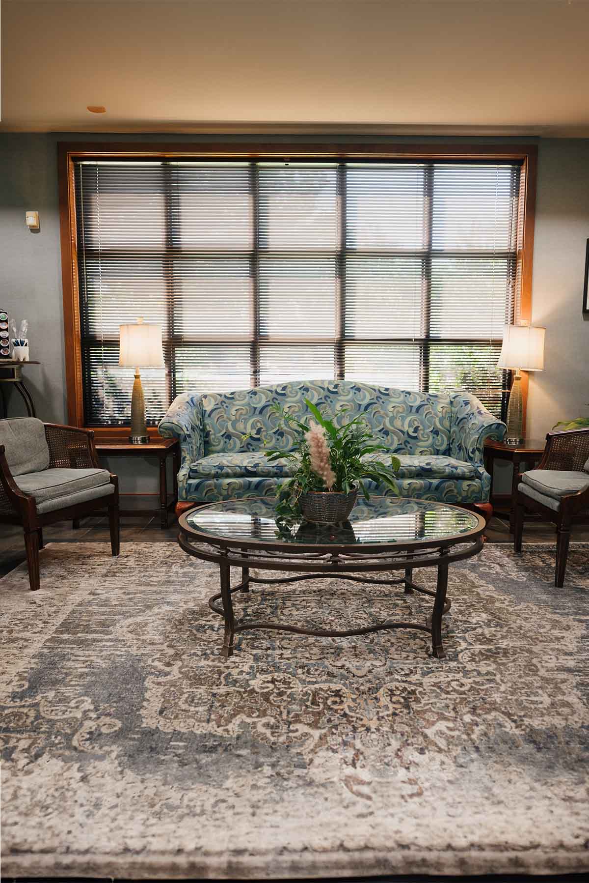 Living room with a patterned blue sofa, two gray cushioned chairs, a glass coffee table with a green plant, and large window blinds.