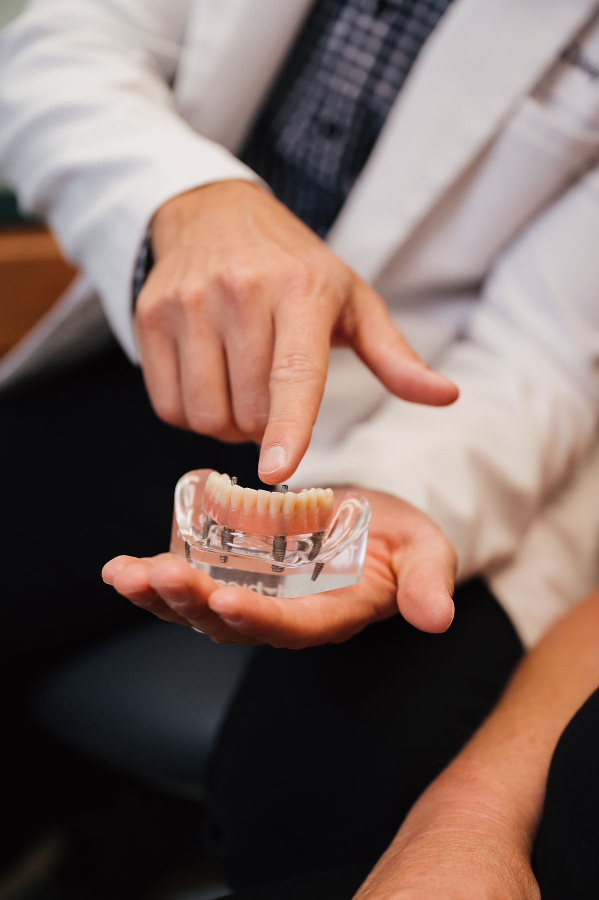 Person in a white coat holding and pointing at a dental implant model with artificial teeth.