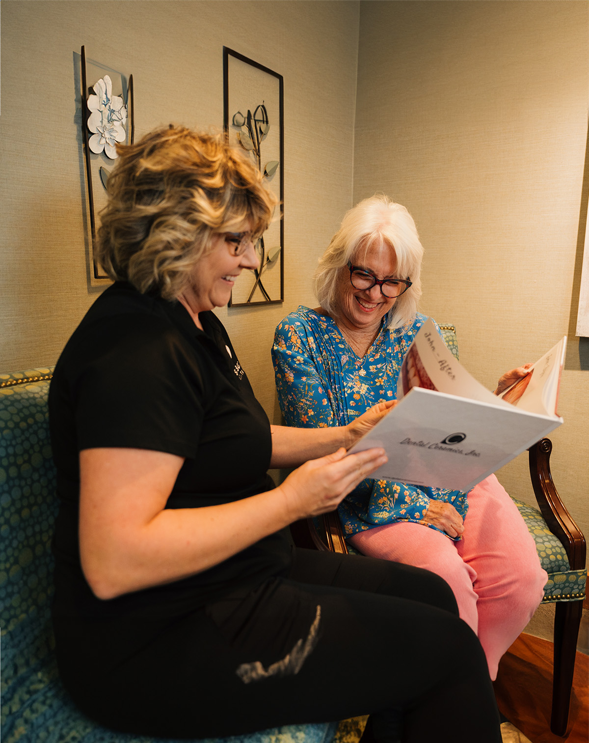 Two women sitting and smiling while looking at a large book in a cozy room.