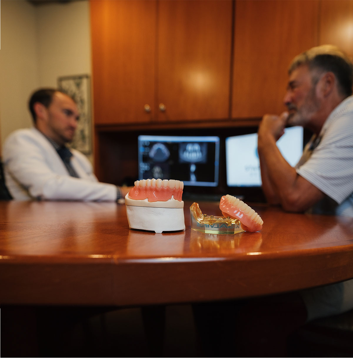Dental prosthetics on a wooden table with two men, one in a white coat, consulting in the background with dental images on monitors.
