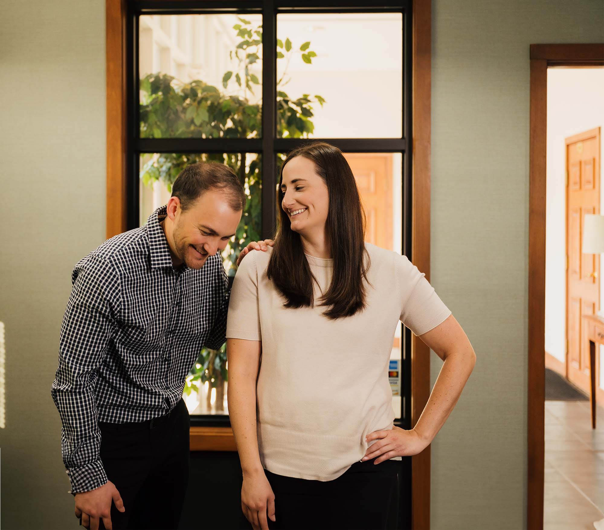 A woman in a beige top smiling and resting her hand on the shoulder of a man in a checkered shirt who is laughing and looking down.
