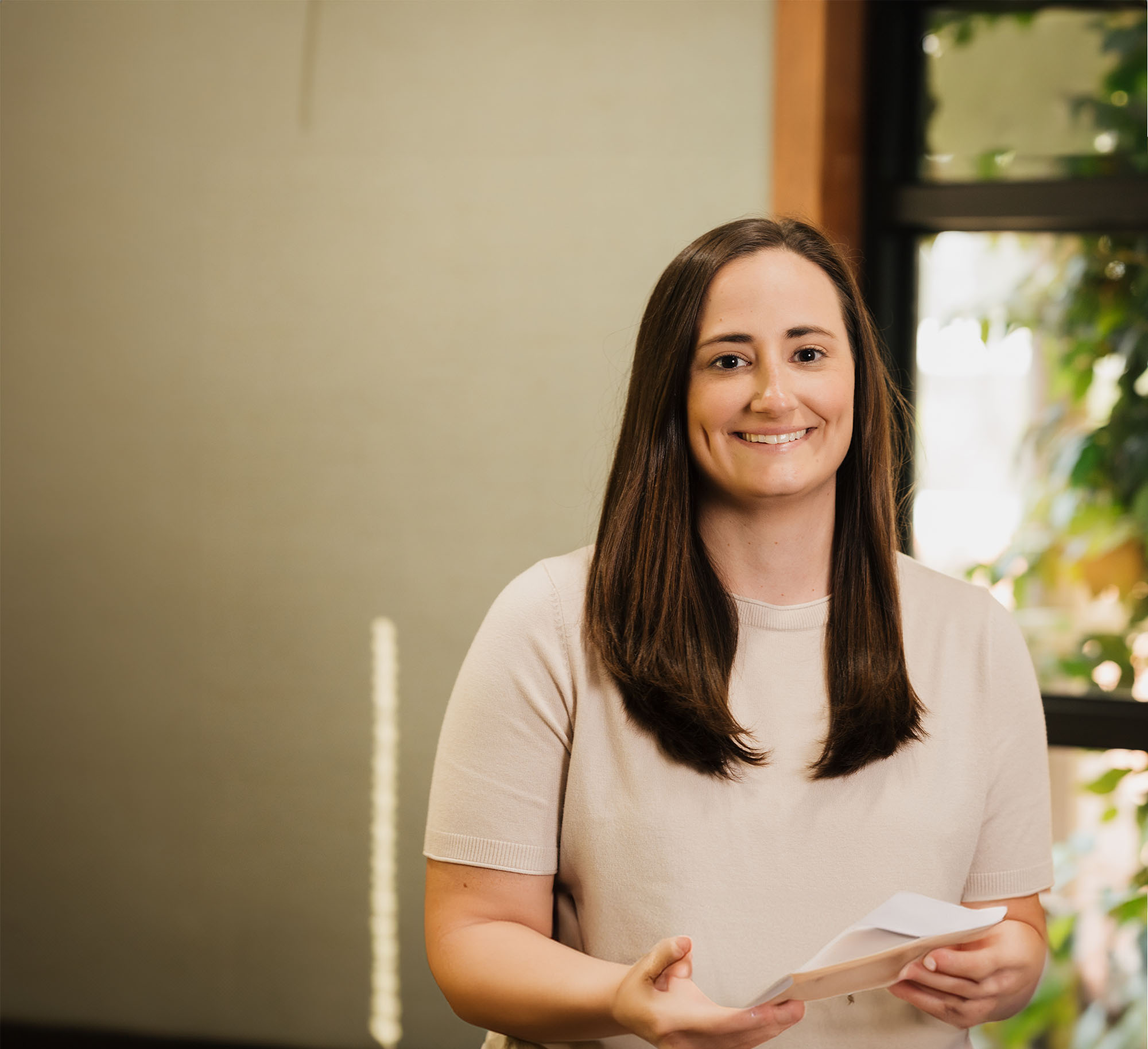 Smiling woman with long brown hair holding folded papers in a softly lit room with a window and plants in the background.