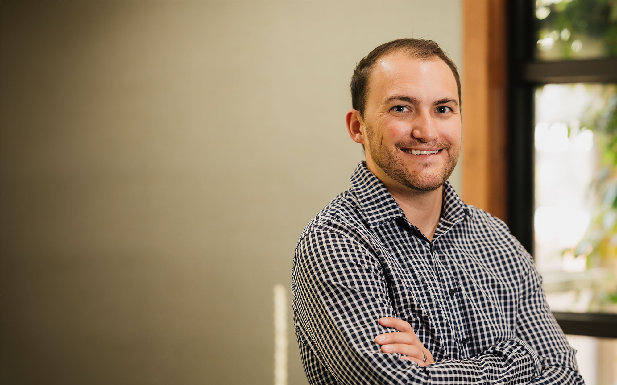 Smiling man in a black-and-white checkered shirt standing with arms crossed indoors near a window.