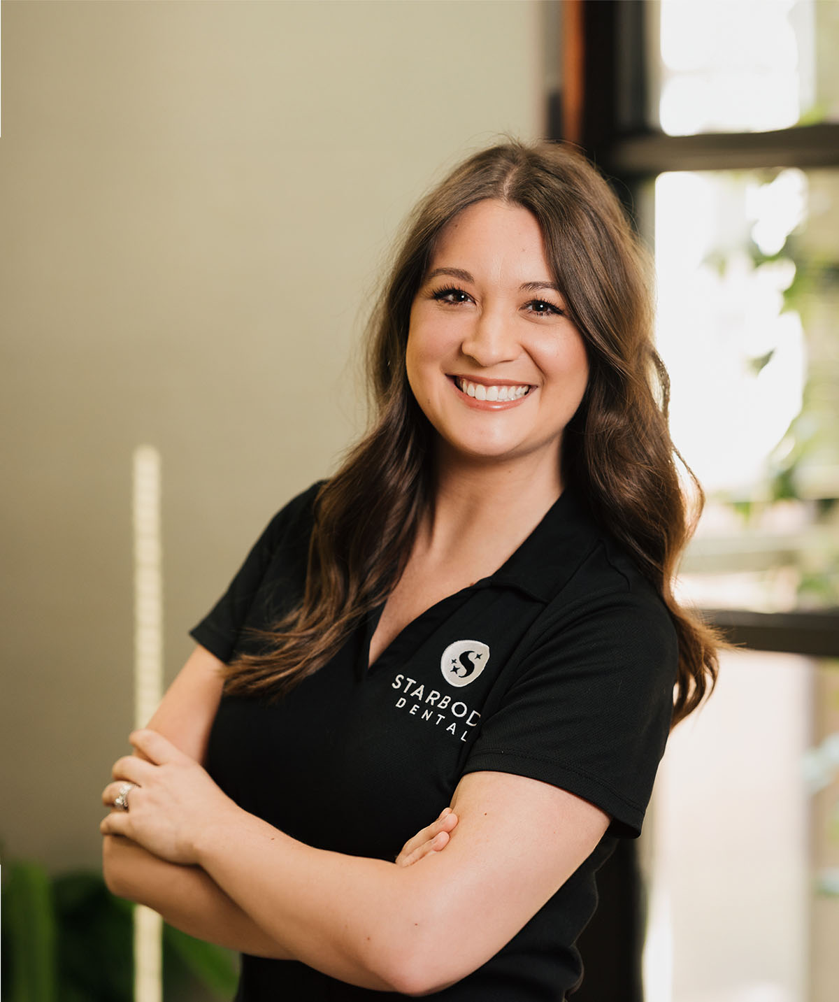 Smiling woman with long brown hair wearing a black Starbody Dental polo shirt standing with arms crossed indoors.