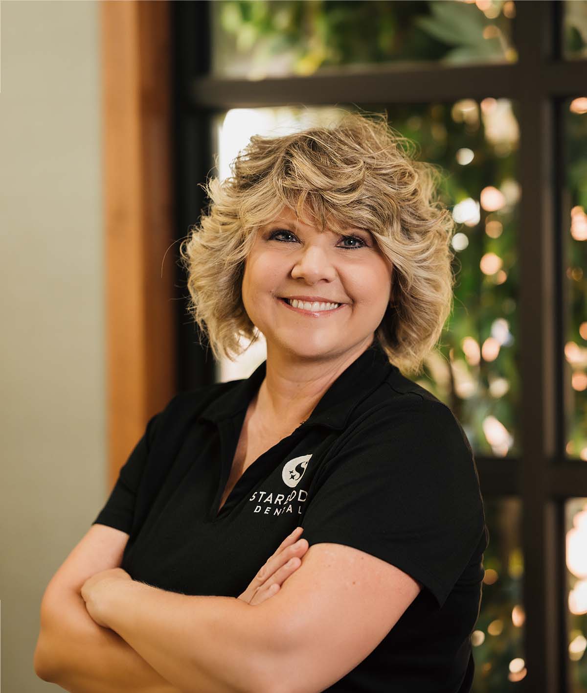 Smiling woman with short curly blonde hair wearing a black Starbody Dental polo shirt, standing with arms crossed indoors.