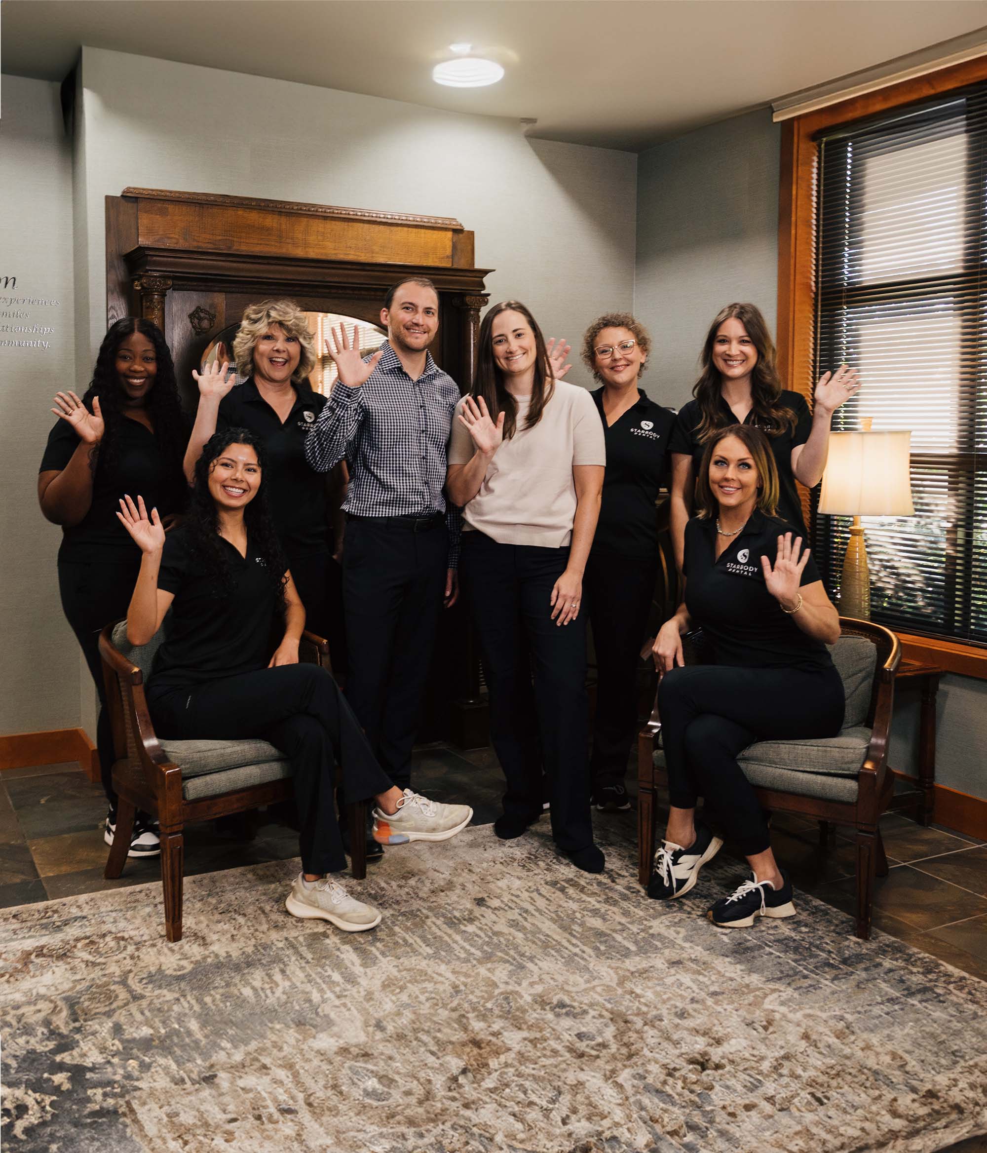 Group of eight diverse dental office staff smiling and waving indoors with two seated in chairs and six standing behind.