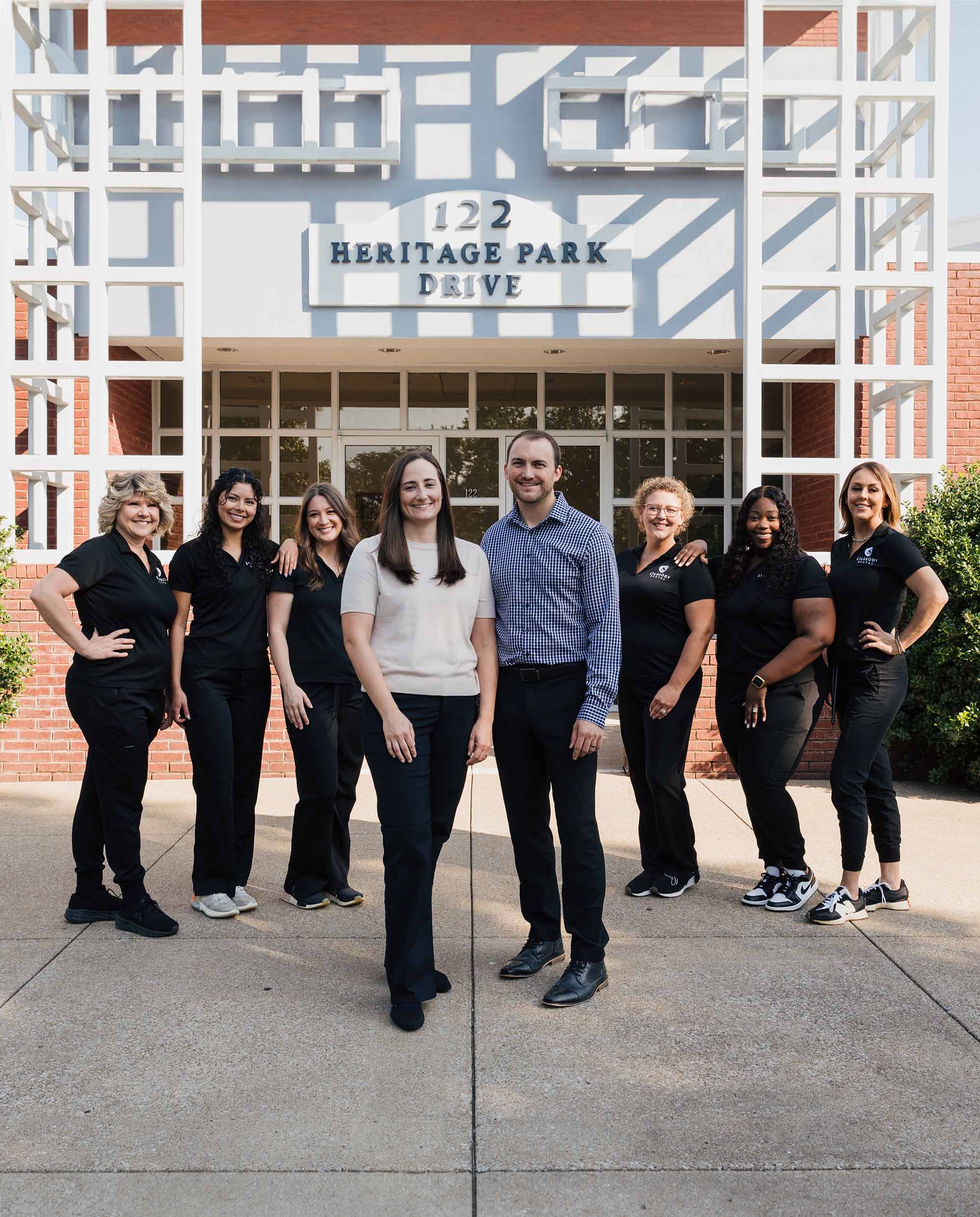 Group of eight diverse dental professionals standing outside a building with the address 122 Heritage Park Drive, smiling at the camera.