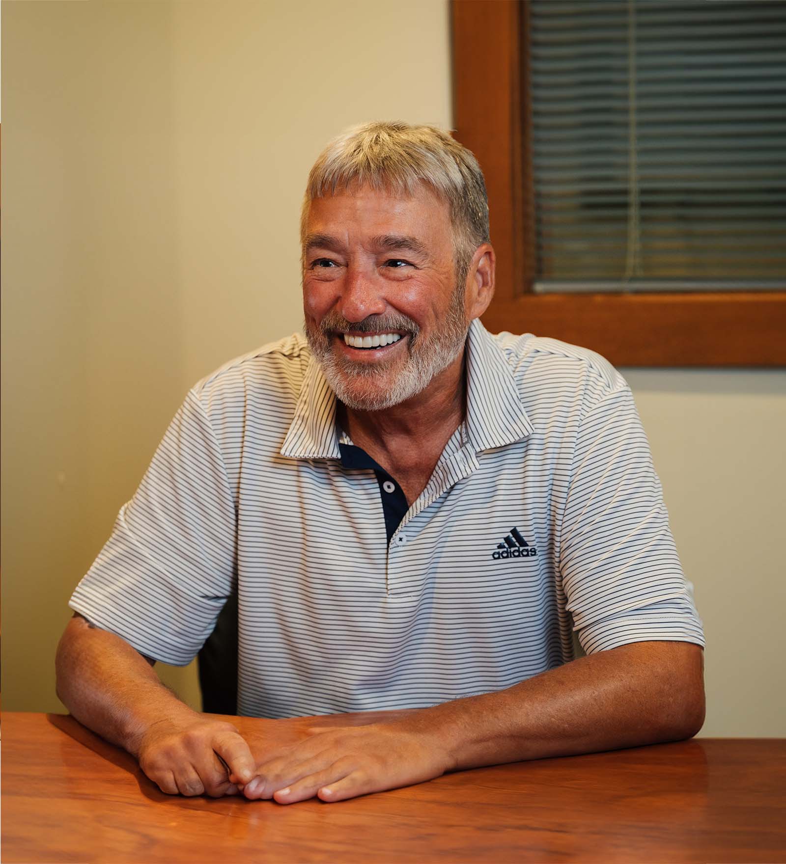 Smiling middle-aged man with gray hair and beard wearing a white Adidas striped polo shirt sitting at a wooden table.