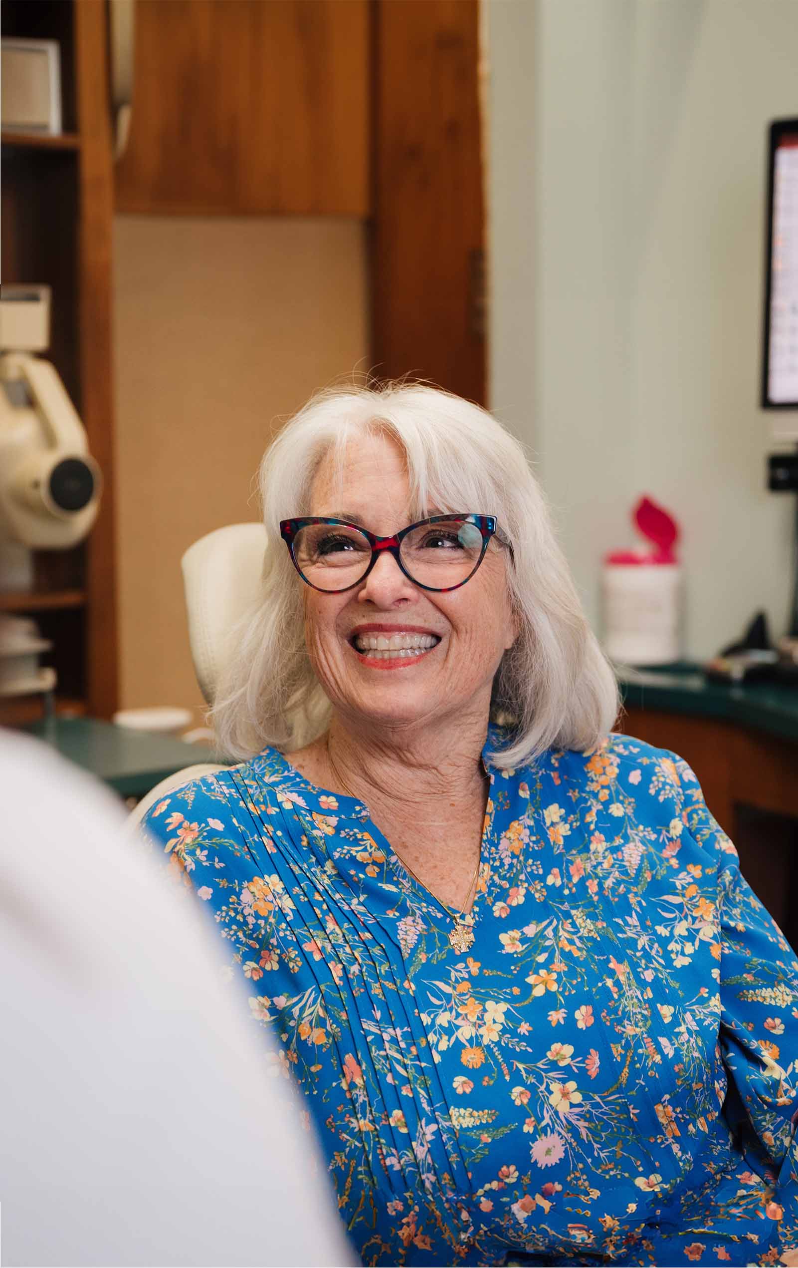 Smiling elderly woman with white hair and glasses sitting in a dental office chair wearing a blue floral blouse.