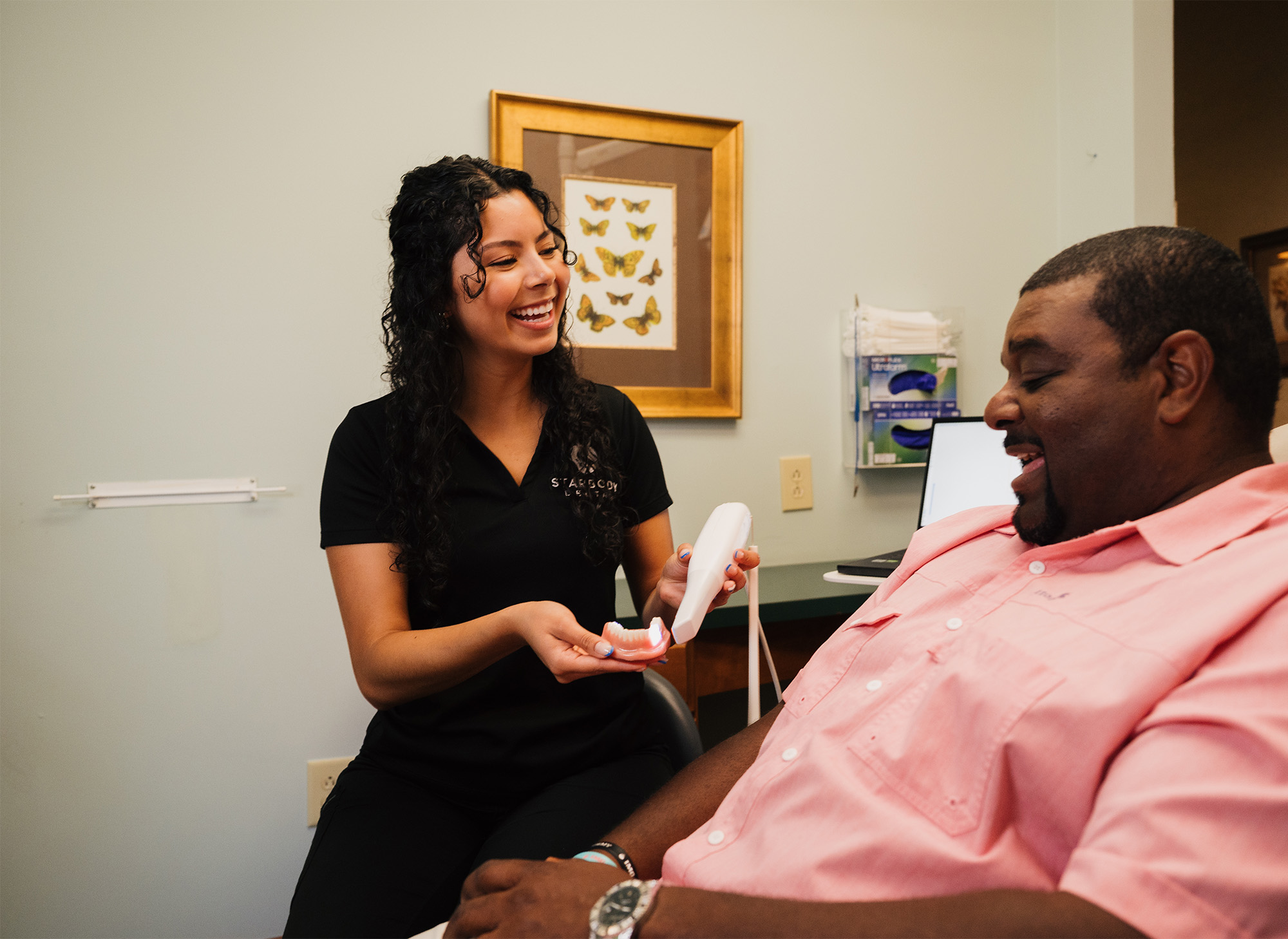 Female dental professional smiling and holding a dental scanner and model while interacting with a seated male patient in a dental office.